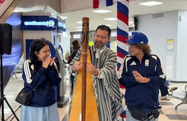 Un hombre toca el arpa vestido con ropa tradicional, mientras una mujer sonriente y un hombre aplauden con chaquetas deportivas en un ambiente festivo.