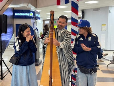 Un hombre toca el arpa vestido con ropa tradicional, mientras una mujer sonriente y un hombre aplauden con chaquetas deportivas en un ambiente festivo.