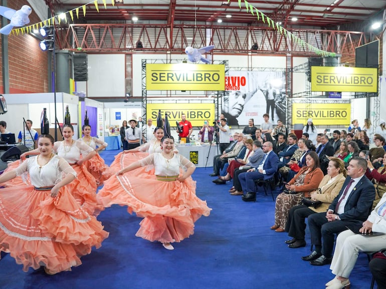 Seis mujeres en trajes tradicionales naranjas y blusas blancas bailan en un evento festivo, rodeadas de una audiencia diversa.