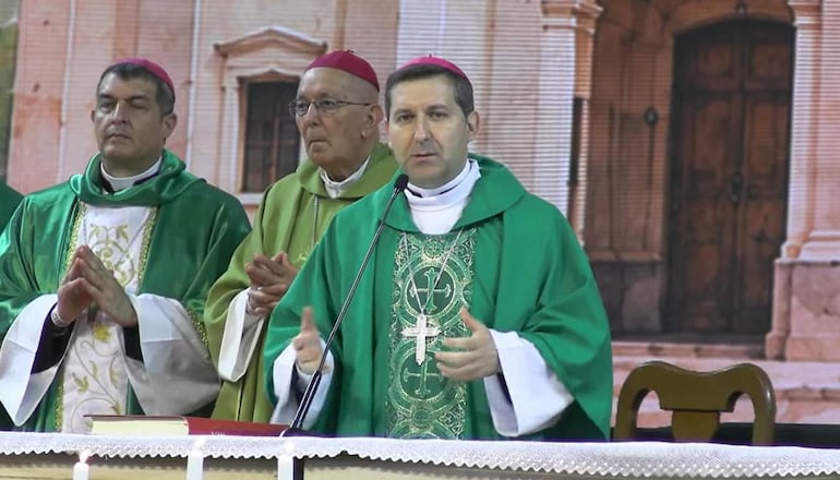 Hombres con túnicas verdes y doradas, orador en micrófono, ambiente religioso con columnas y puertas visibles.