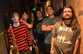 Músicos de Vecindad Autopsia posan en backstage. Hombre con saxofón y camiseta de rayas, cuatro compañeros con diversas camisetas.
