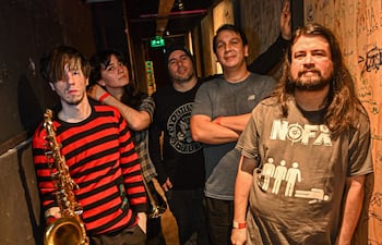 Músicos de Vecindad Autopsia posan en backstage. Hombre con saxofón y camiseta de rayas, cuatro compañeros con diversas camisetas.