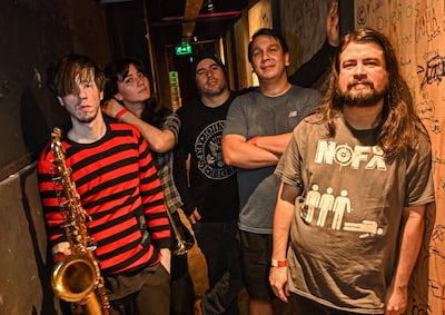 Músicos de Vecindad Autopsia posan en backstage. Hombre con saxofón y camiseta de rayas, cuatro compañeros con diversas camisetas.
