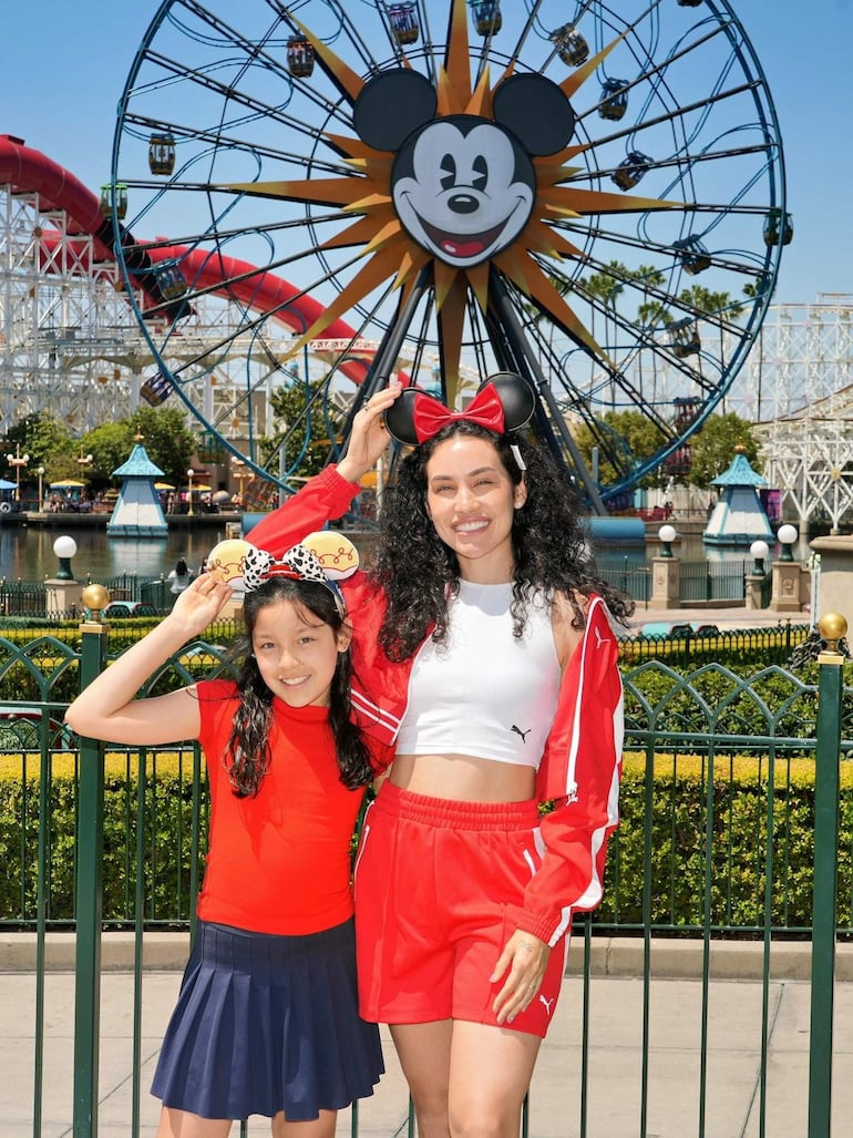 Mujer adulta con parte superior blanca y pantalones cortos rojos, y niña en camiseta roja y falda azul, sonríen en un parque de diversiones.