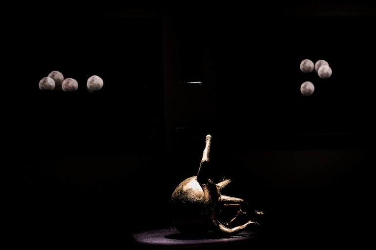 Escultura dorada en primer plano con fondo oscuro, destacándose en un ambiente de galería de arte sin presencia de personas.