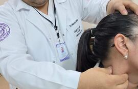 Médico en uniforme blanco examina el cuello de una mujer con abrigo claro, ambos en un consultorio médico.