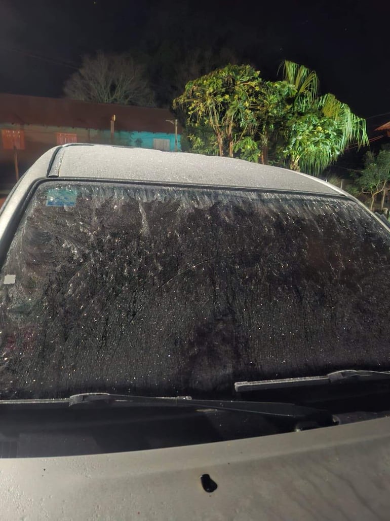 Vehículo estacionado con parabrisas húmedo, rodeado de naturaleza y estructura de colores variados en ambiente nocturno.