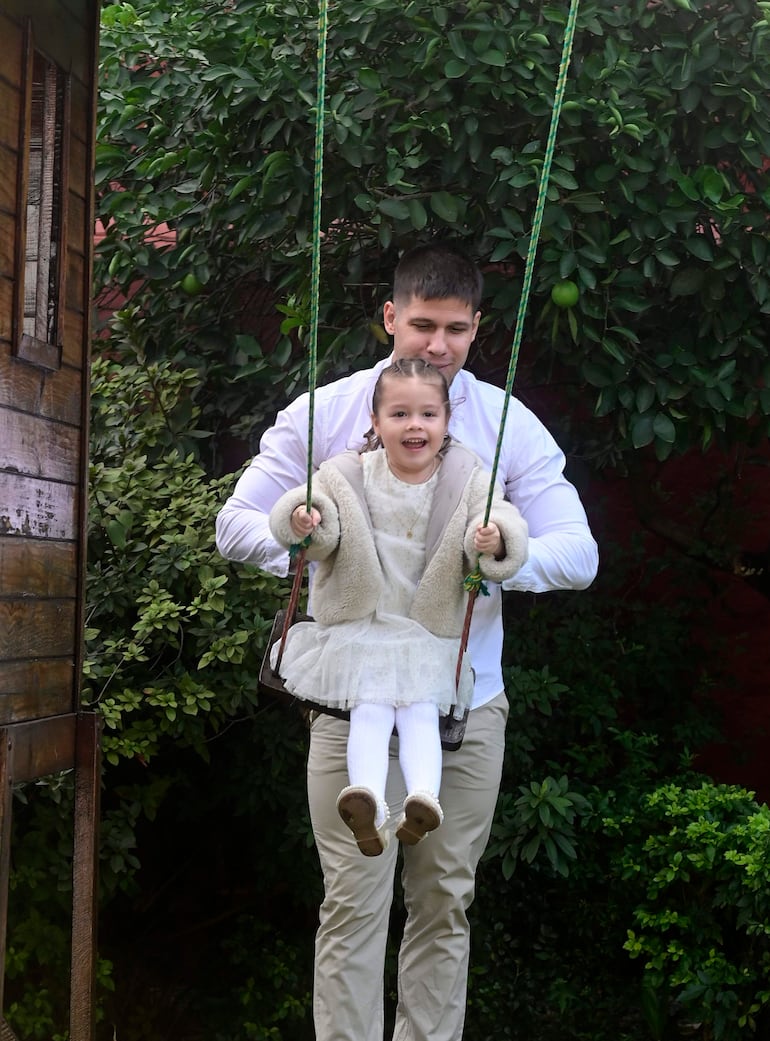 Charles Hockin en camisa blanca sostiene un columpio donde ríe su hija con vestido blanco en un entorno verde.