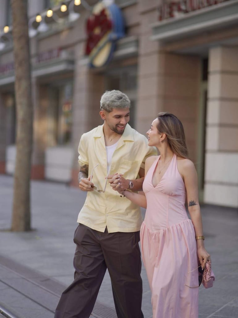 Julio Enciso con camisa clara y gafas en mano, junto a Ramón Sosa, quien lleva vestido rosa, ambos sonrientes en la calle.