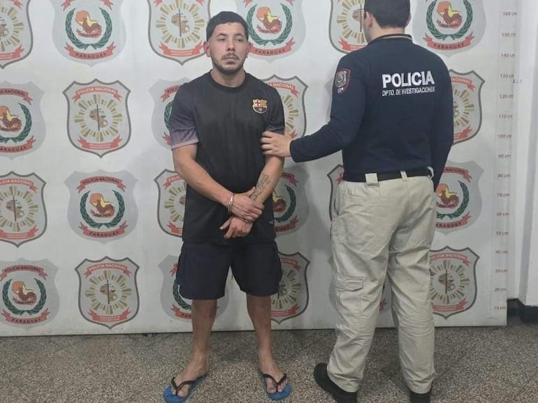 Hombre de pie en camiseta oscura con logo deportivo, custodiado por un policía en uniforme, en ambiente tenso de instalación policial.