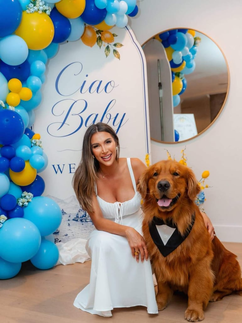 Mujer con cabello suelto y vestido blanco sonríe junto a un perro Retriever dorado con moño negro, en un entorno festivo.