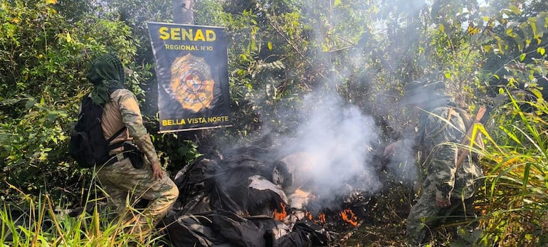Dos agentes antidrogas vestidos con atuendos militares junto a un fuego, en una operación de erradicación de drogas en la vegetación densa.