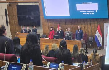 Mujer en abrigo rojo destaca en podio junto a hombres de traje, en un recinto legislativo moderno con banderas paraguayas.