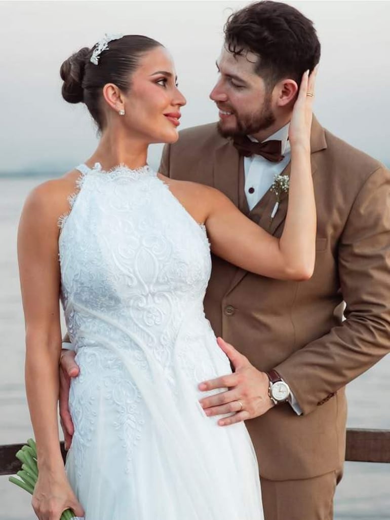 Hombre en traje marrón claro y mujer en vestido blanco se miran con cariño frente a un lago en un ambiente festivo.