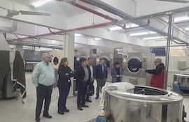 Hombre mayor en abrigo rojo gesticula mientras explica a un grupo en una lavandería industrial con máquinas visibles al fondo.