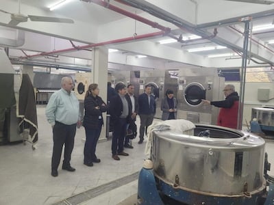 Hombre mayor en abrigo rojo gesticula mientras explica a un grupo en una lavandería industrial con máquinas visibles al fondo.