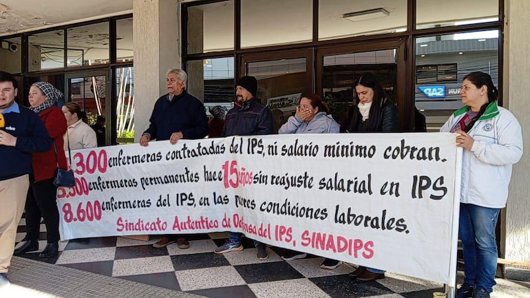 Grupo de enfermeras con expresión de preocupación sostiene un cartel frente a un edificio, mientras un hombre habla al micrófono.