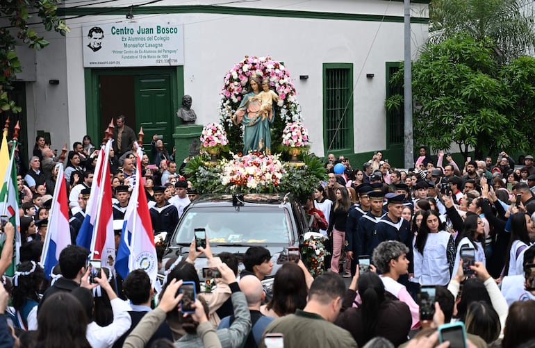 Niños en uniformes y asistentes sosteniendo banderas durante la procesión, con un vehículo decorado como fondo.