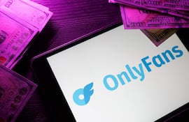 Tablet con logotipo de OnlyFans en pantalla, rodeado de billetes de $100 en un ambiente interior y bien iluminado.