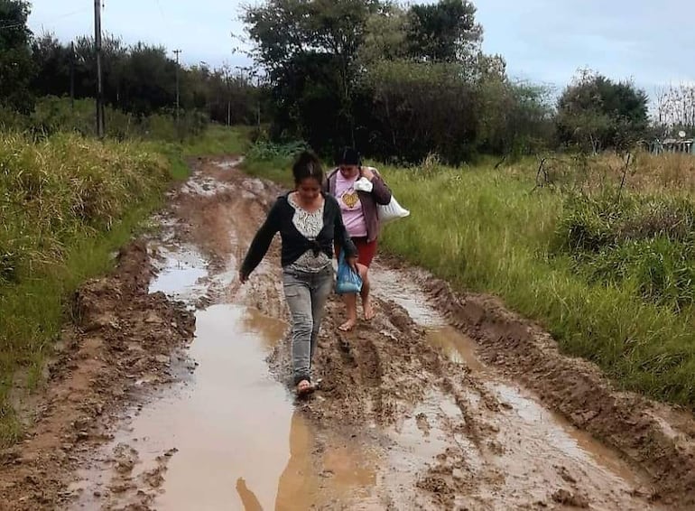 Dos mujeres caminan por un camino embarrado; una viste abrigo negro y jeans claros, la otra lleva camiseta rosa y bolsa blanca.