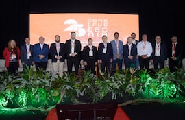 Grupo de diez personas vestidas formalmente frente a un escenario con cartel del 25° Construcción, en ambiente decorado con plantas.