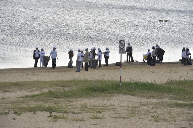 Grupo de personas vestidas con camisetas blancas limpian la orilla, llevando bolsas de basura en un entorno natural.