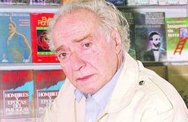 Elvio Romero, hombre de edad avanzada con cabello canoso, sentado en una biblioteca, con una expresión seria y rodeado de estantes de libros.