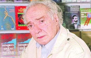 Elvio Romero, hombre de edad avanzada con cabello canoso, sentado en una biblioteca, con una expresión seria y rodeado de estantes de libros.