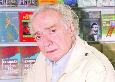 Elvio Romero, hombre de edad avanzada con cabello canoso, sentado en una biblioteca, con una expresión seria y rodeado de estantes de libros.