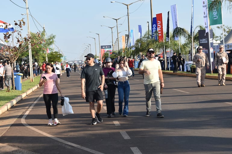 Mujer con teléfono y hombre a su lado caminan; soldados en uniforme militar están de pie en el fondo, rodeados de banderas.