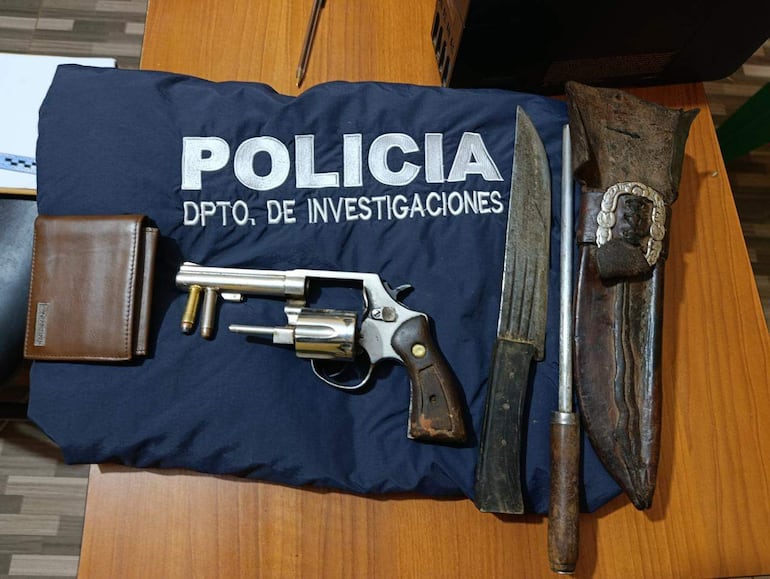 Chaqueta policial con bordado 'POLICÍA', revólver y otros objetos dispuestos sobre mesa de madera en ambiente interior.