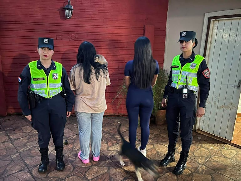 Tres policías con chalecos de alta visibilidad y una mujer sosteniendo un mono en un ambiente exterior frente a un muro rojo.