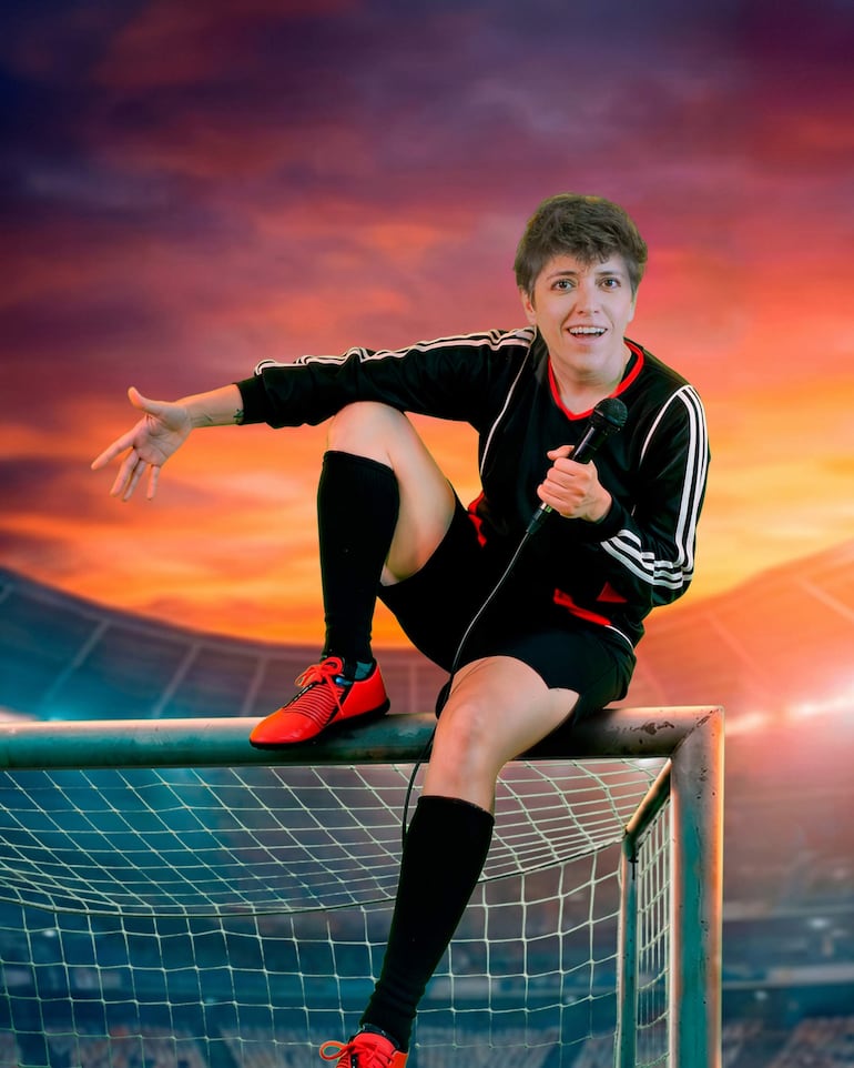 Caro Romero en uniforme deportivo negro y rojo, con micrófono en mano, gesticulando en el travesaño de una portería al atardecer.