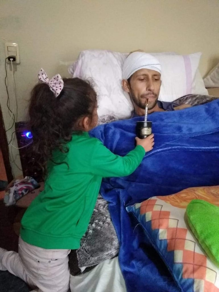 Niña de cabello rizado en sudadera verde ofrece mate a hombre con venda en la cabeza en un ambiente acogedor y familiar.
