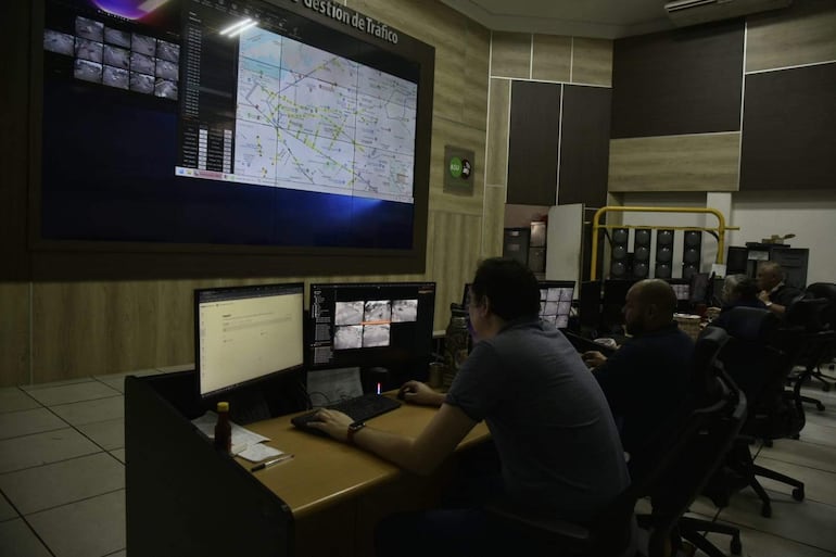 Hombres en un centro de control de tráfico, trabajando en computadoras con pantallas de monitoreo y un mapa en el fondo.
