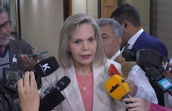 Lilian Samaniego en blazer claro y blusa roja, rodeada de periodistas que la entrevistan en un ambiente institucional.