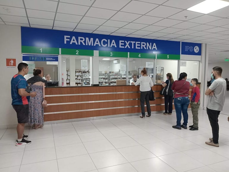 Personas con mascarillas en farmacia, hombres y mujeres conversan, ambiente moderno. Una mujer de vestido largo y un hombre de camiseta azul en primer plano.