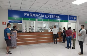 Personas con mascarillas en farmacia, hombres y mujeres conversan, ambiente moderno. Una mujer de vestido largo y un hombre de camiseta azul en primer plano.