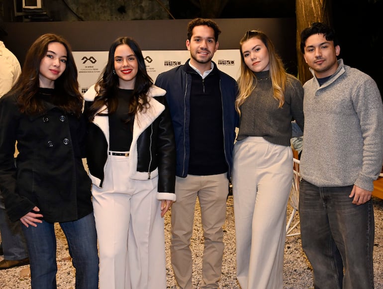 Cinco personas sonríen posando: tres mujeres y dos hombres con ropa de abrigo, en un evento festivo decorado.