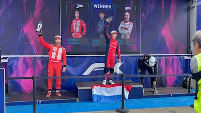 Mateo Valente en traje rojo sostiene un trofeo, sonriente. Valentino Carofano y Felisa Blanco lo acompañan en el podio con expresiones variadas.