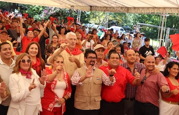 Arnoldo Wiens sonríe al centro, rodeado de personas con ropa roja y blanca, gesticulando en un evento al aire libre.