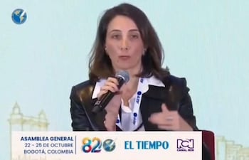 Mujer con chaqueta negra y blusa blanca sostiene un micrófono, hablando en un auditorio decorado con banners del evento.