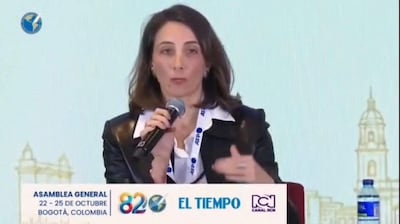 Mujer con chaqueta negra y blusa blanca sostiene un micrófono, hablando en un auditorio decorado con banners del evento.