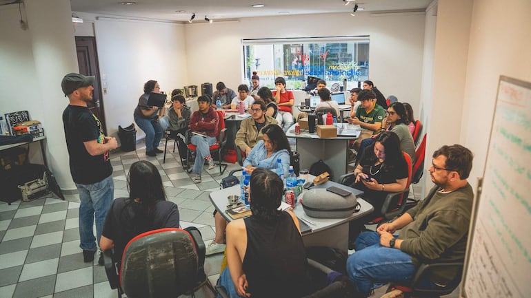 Hombre con gorra y camiseta negra presenta en sala, rodeado de asistentes que toman notas y observan atentos.