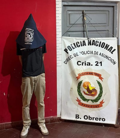 Joven con capucho oscuro de pie en comisaría, viste camiseta negra y pantalones claros, junto a cartel de Policía Nacional.