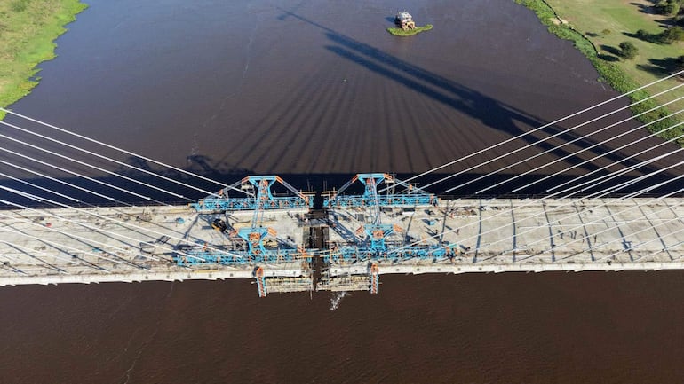 Vista aérea del puente en construcción con cables azules y naranjas, rodeado de vegetación y un bote trabajando en el agua.
