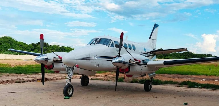 Avión Beechcraft blanco y negro estacionado en área desierta, rodeado de vegetación y un cielo despejado.