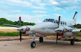 Avión Beechcraft blanco y negro estacionado en área desierta, rodeado de vegetación y un cielo despejado.