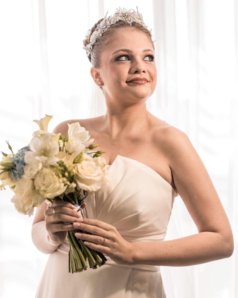 Mujer en vestido blanco sin mangas y tiara, sonríe mientras sostiene un ramo de flores blancas y azules en un entorno iluminado.