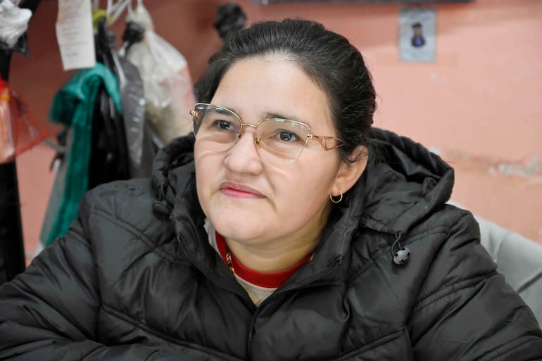 Mujer con cabello oscuro recogido, gafas y chaqueta negra, sentada en un entorno con elementos personales poco definidos.